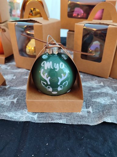 Boule de Noël verte ornée de motifs blancs et portant le nom "Mya", présentée dans un emballage.